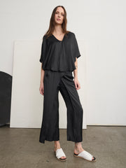 A woman poses confidently on a concrete floor in a minimalist room, wearing lightweight black Ido Pants with an elastic waist by Zero + Maria Cornejo and white sandals. Her long hair complements the simple backdrop.