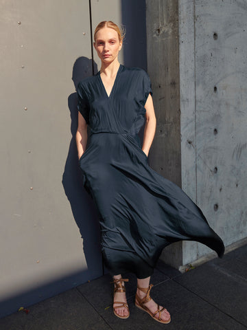 In a sunlit scene, a person stands against a concrete wall wearing Zero + Maria Cornejo's Long Aki Wave Dress, known for its dark, lightweight georgette fabric with an asymmetrical skirt, paired with sandals. The dress catches the wind, creating dynamic shadows on the wall.