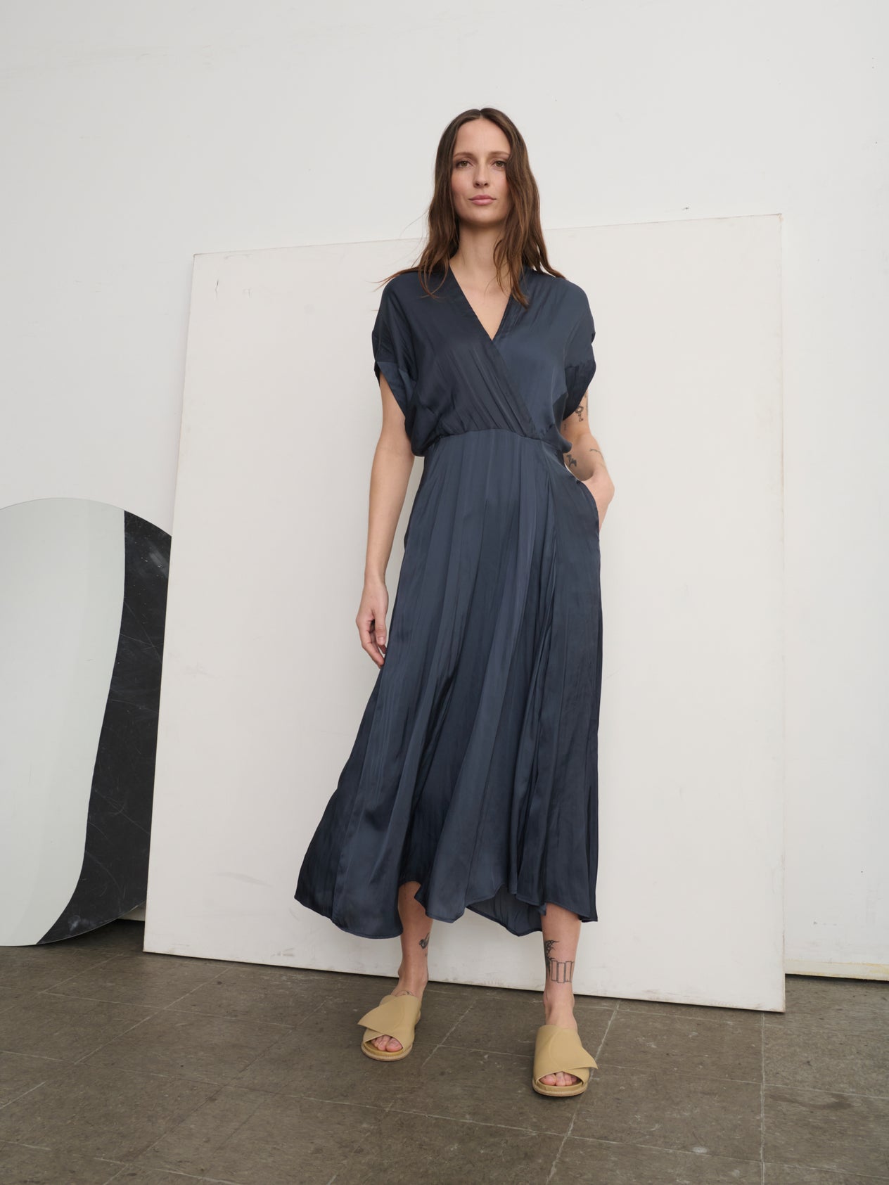 Against a plain backdrop, a person with long hair wears beige slide sandals and the Zero + Maria Cornejo Long Aki Wave Dress, featuring a wrap-effect V-neck and subtle asymmetrical skirt. With hands in pockets, they exude an elegant yet casual vibe in this flowing georgette dress.