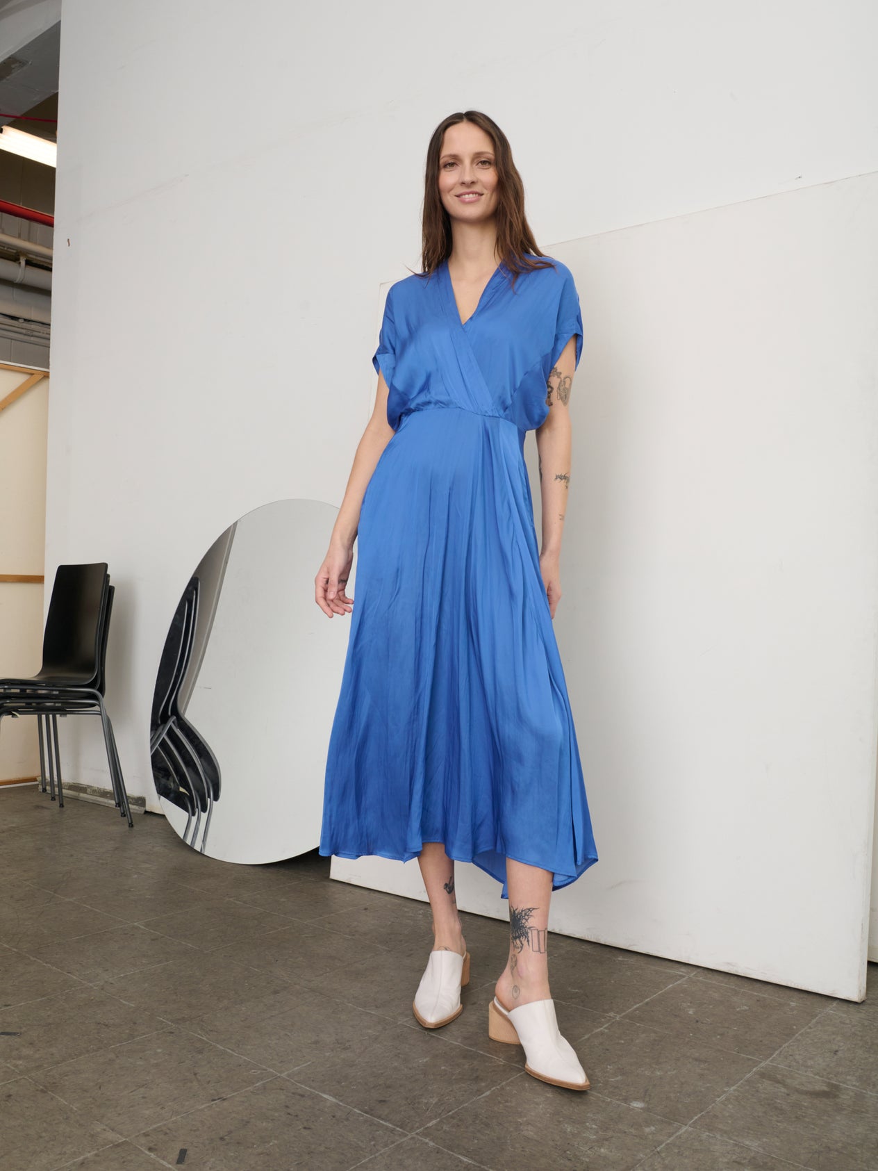 Against a plain backdrop, a person with long hair wears beige slide sandals and the Zero + Maria Cornejo Long Aki Wave Dress, featuring a wrap-effect V-neck and subtle asymmetrical skirt. With hands in pockets, they exude an elegant yet casual vibe in this flowing georgette dress.
