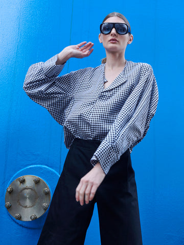 In front of a bright blue background with a metal fitting, a person poses confidently wearing oversized sunglasses, black pants, and the Long-Sleeved Akeo Shirt by Zero + Maria Cornejo.