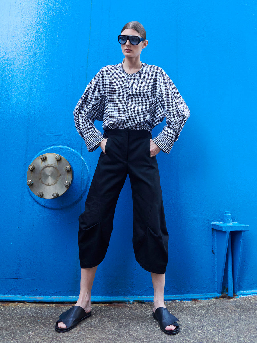 A person confidently stands against a bright blue wall, wearing sleek black sunglasses, a Long-Sleeved Akeo Shirt by Zero + Maria Cornejo, and black pants. Black slide sandals complete the look as they stand with hands on hips.