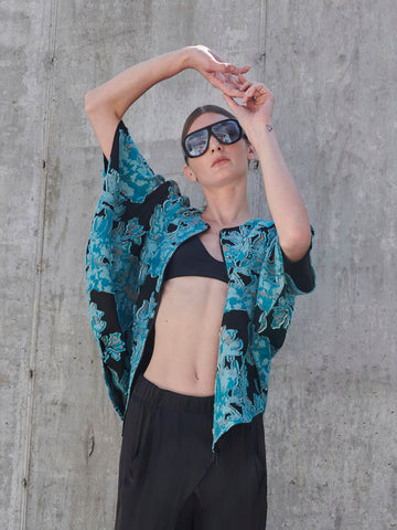 A person poses against a concrete wall wearing large sunglasses and the Zero + Maria Cornejo Circle Shrug, a blue organic floral jacquard shirt draped over a black top. Dark pants complete the look as they gracefully raise their arms above their head.