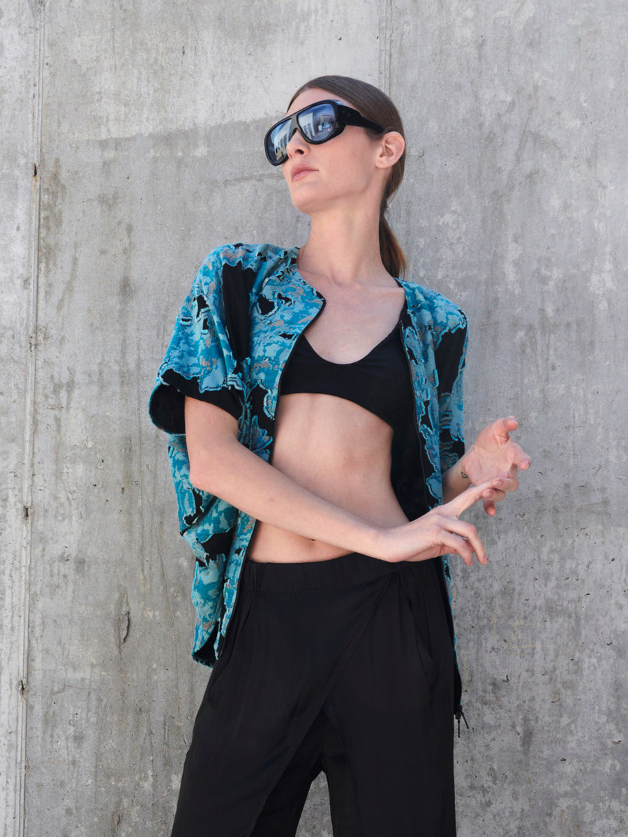 A person stands against a concrete wall, exuding the draped silhouette style in dark sunglasses, a black crop top, and pants. They add flair with an open Hawaiian-style shirt featuring blue organic floral jacquard patterns. One arm is relaxed, the other elegantly bent. Brand: Zero + Maria Cornejo's Circle Shrug.