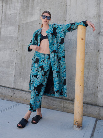 A person poses against a concrete wall, wearing sunglasses and the Eve Koya Coat from Zero + Maria Cornejo, featuring an organic cotton teal floral-patterned matching set with pants and a black top. They sport black sandals and lean casually against a yellow pole.