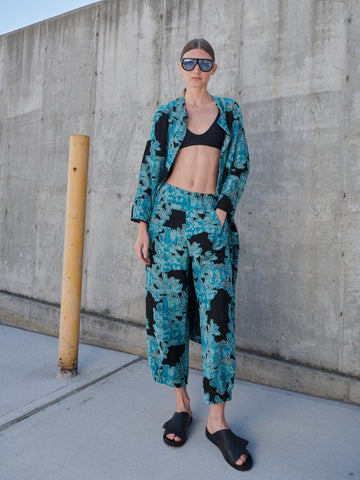 A person stands against a concrete wall in the Abi Pant by Zero + Maria Cornejo, featuring a blue floral jacquard design. Paired with a black crop top, slide sandals, sunglasses, and tied-back hair, this stylish look highlights sustainably-sourced materials for a conscious fashion statement.