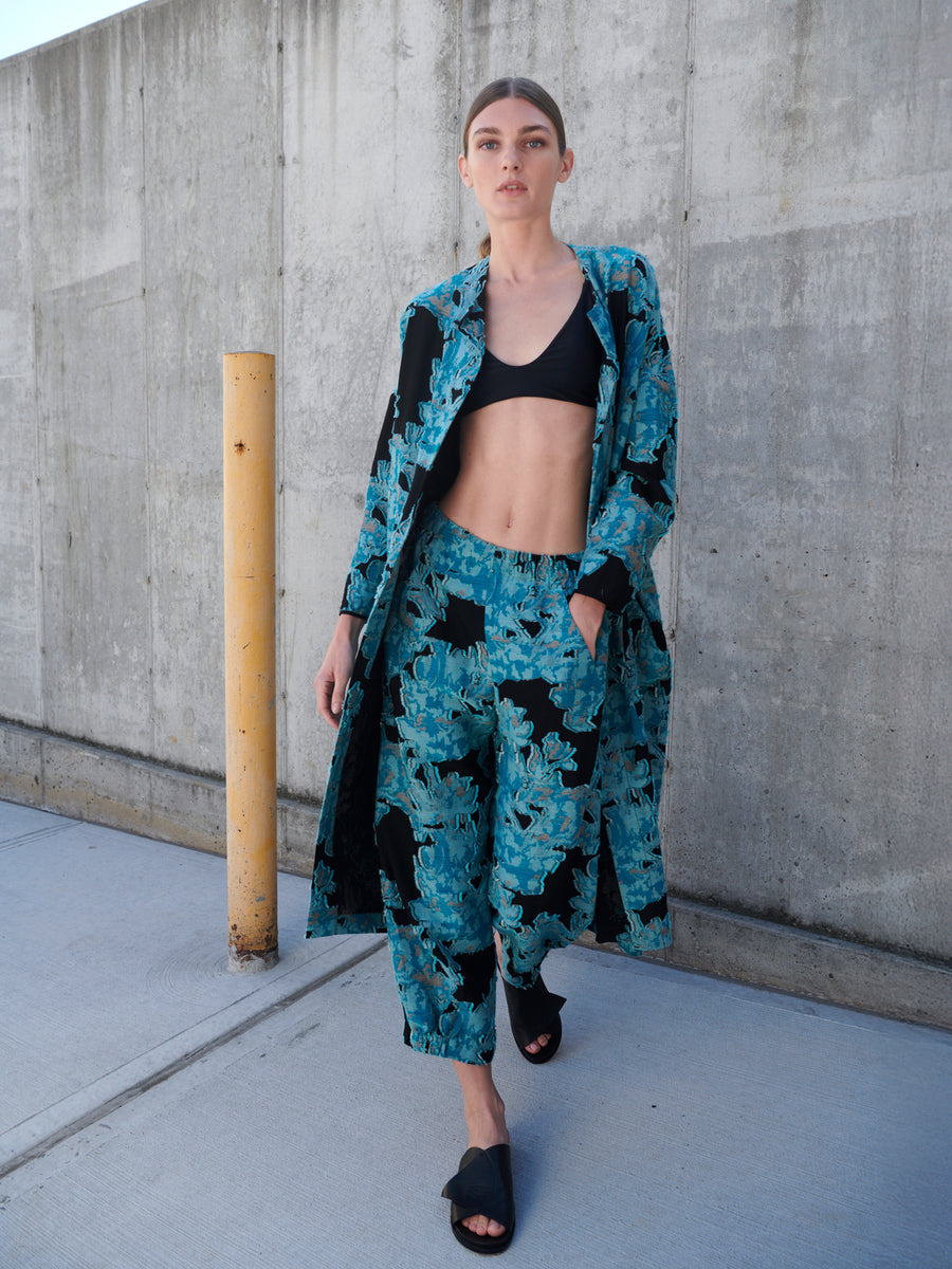 A woman poses against a concrete wall in the Abi Pant by Zero + Maria Cornejo, a black crop top, and teal floral jacquard pantsuit. Her hair is pulled back, and she's wearing black sandals. A yellow post complements her stylish look made from sustainably-sourced materials.