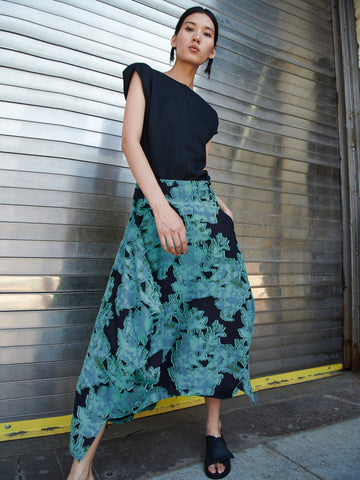 A person confidently poses against a metallic, corrugated backdrop, wearing a sleeveless black top, the Nio Skirt by Zero + Maria Cornejo with a green floral pattern, and black sandals on a concrete surface.