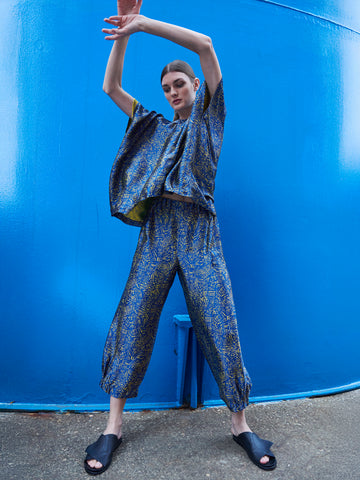 Posing stylishly against a bright blue wall, they don the Abi Pant from Zero + Maria Cornejo, a loose-fitting blue outfit made with FSC certified viscose, paired with black slide sandals. Their raised arms highlight the vibrant colors and sustainable materials.