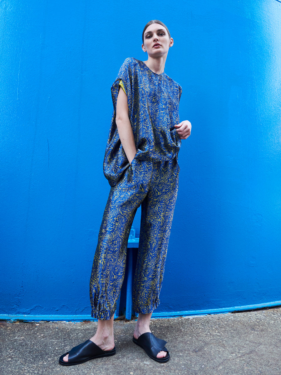 A person stands confidently against a bright blue wall, wearing the Abi Pant by Zero + Maria Cornejo, a loose-fitting outfit with abstract designs made from FSC certified viscose. Their hands are in their pockets while they sport black slide sandals.