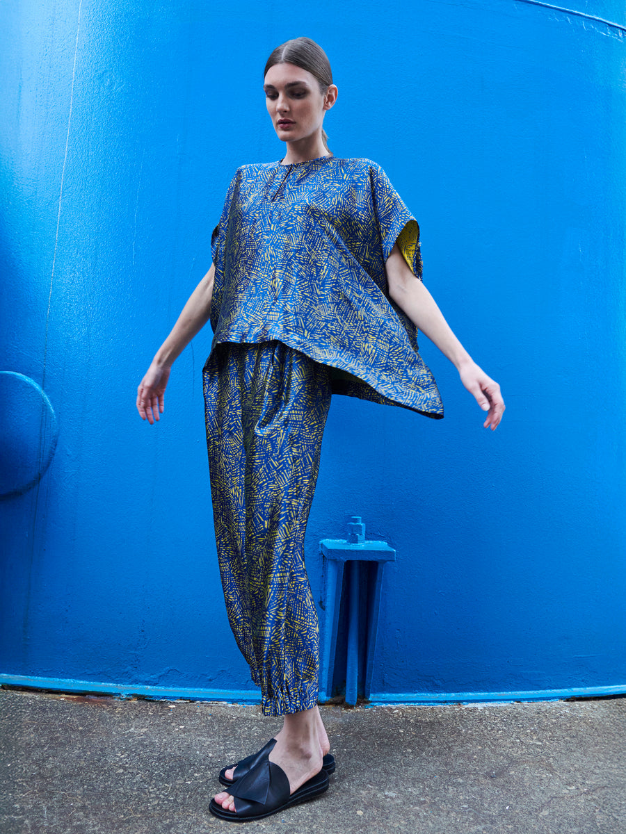 In front of a bright blue wall, a person wears the Abi Pant by Zero + Maria Cornejo, crafted from FSC certified viscose with a blue and yellow abstract pattern. They pair it with black slide sandals, have their hair pulled back, and look downward with arms slightly extended.