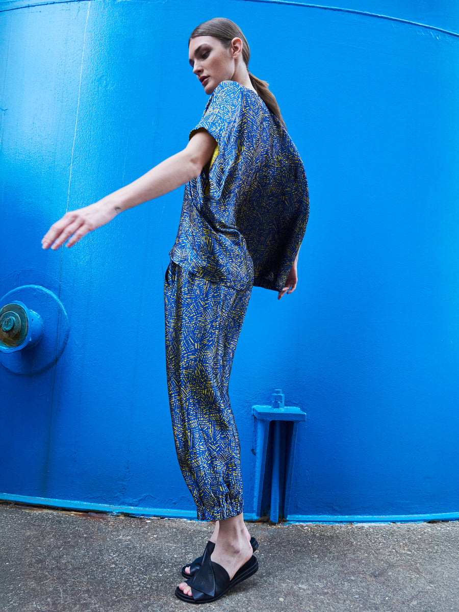 A woman in the Zero + Maria Cornejo Abi Pant, featuring a blue and gold pattern made from FSC certified viscose, stands sideways with one arm extended against a vibrant blue background. She wears black sandals, her hair pulled back, embodying dynamic, fashion-forward elegance through sustainable sourcing.