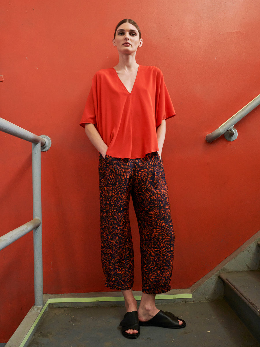 A person is on a staircase against a red wall, wearing a loose red top and Abi Pant by Zero + Maria Cornejo. Their hair is pulled back, and they're in black slip-on shoes. Hands in pockets, they gaze at the camera.