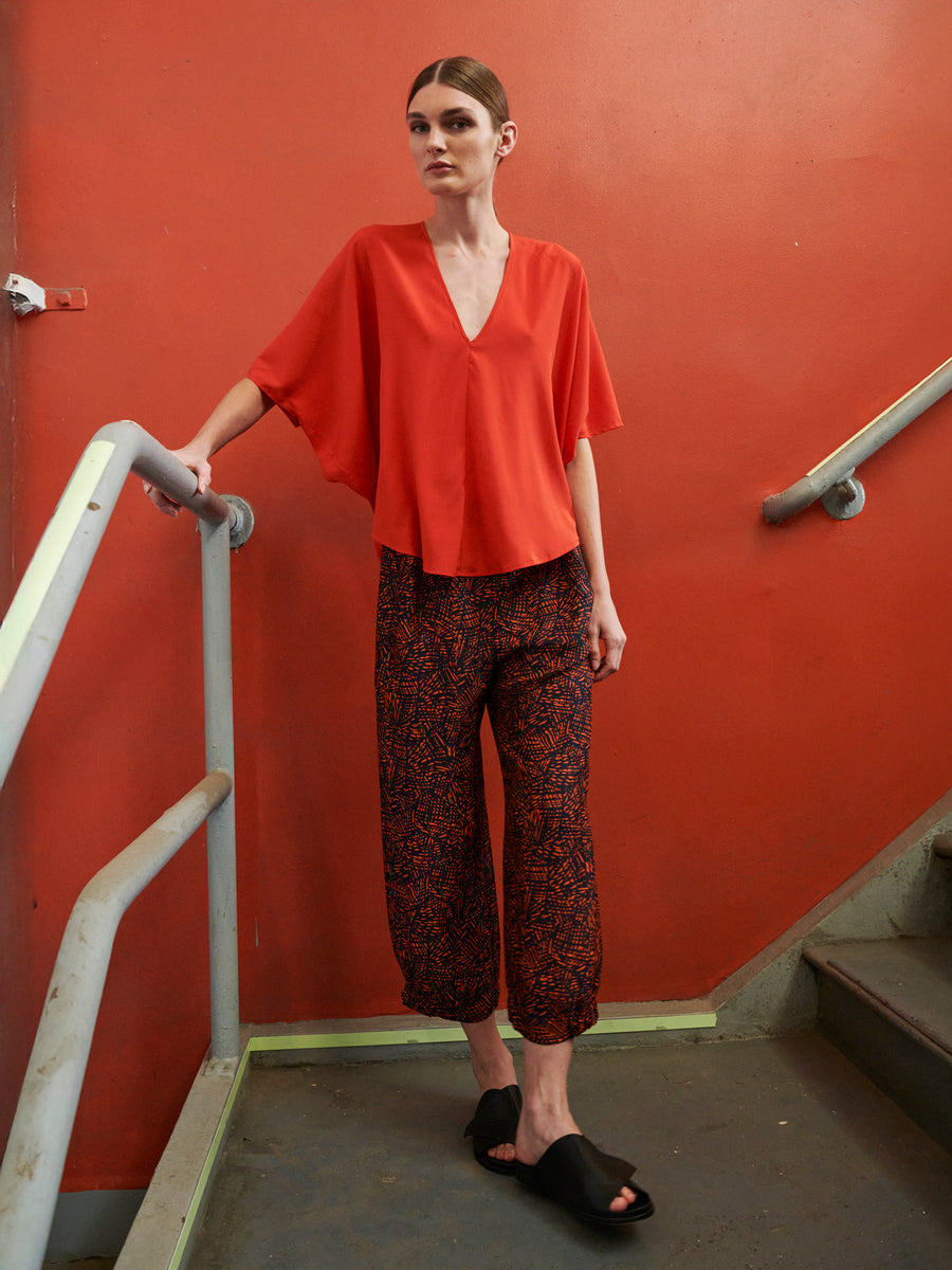 Standing on stairs against a bright red wall, a person wears a loose-fitting red blouse and the Abi Pant by Zero + Maria Cornejo, made from sustainably-sourced materials. They complement their look with black slip-on shoes while gripping the railing.