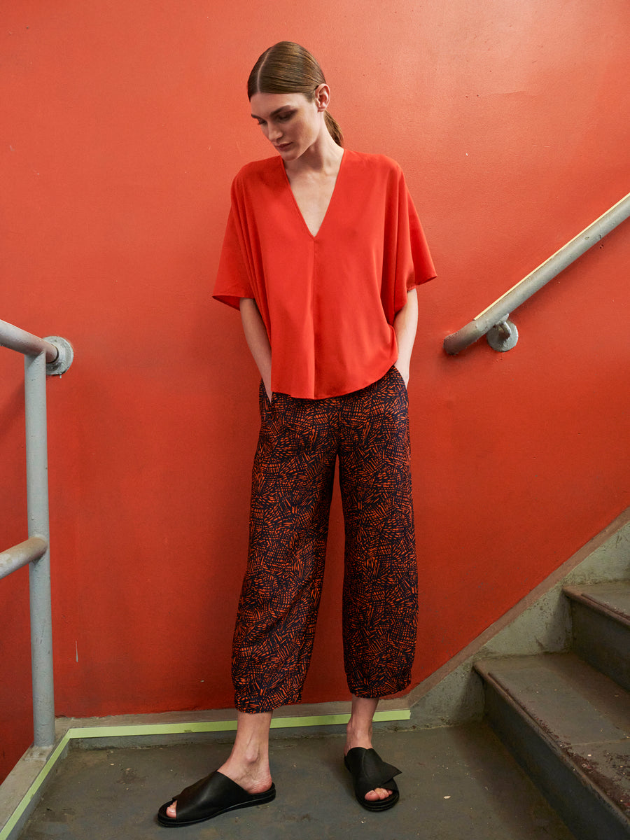 A person stands on a staircase against an orange wall wearing a loose-fitting red V-neck top and graphic jacquard Abi Pants by Zero + Maria Cornejo, styled with black slide sandals. With hair tied back and hands in pockets, they exude relaxed, chic sustainability using FSC-certified viscose fabric.