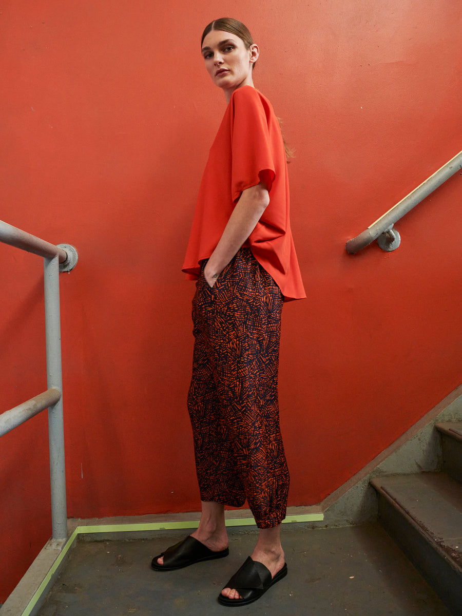 A person stands on a stairwell with orange walls, wearing a bright orange top, Abi Pant by Zero + Maria Cornejo made from FSC certified viscose, and black slide sandals. They have one hand in their pocket and are looking off to the side.