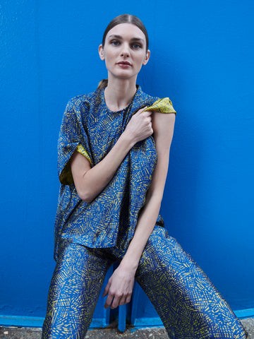 A person with long hair against a bright blue wall wears Zero + Maria Cornejo's Rae Poncho Top in sustainable blue and yellow jacquard, made in New York from FSC certified viscose, holding the fabric at the shoulder with calmness.