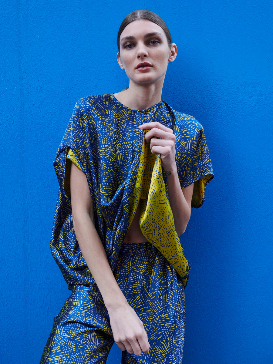 A person with a neutral expression wears the sustainable Rae Poncho Top by Zero + Maria Cornejo against a vibrant blue wall, holding the fabric to highlight its texture and design.