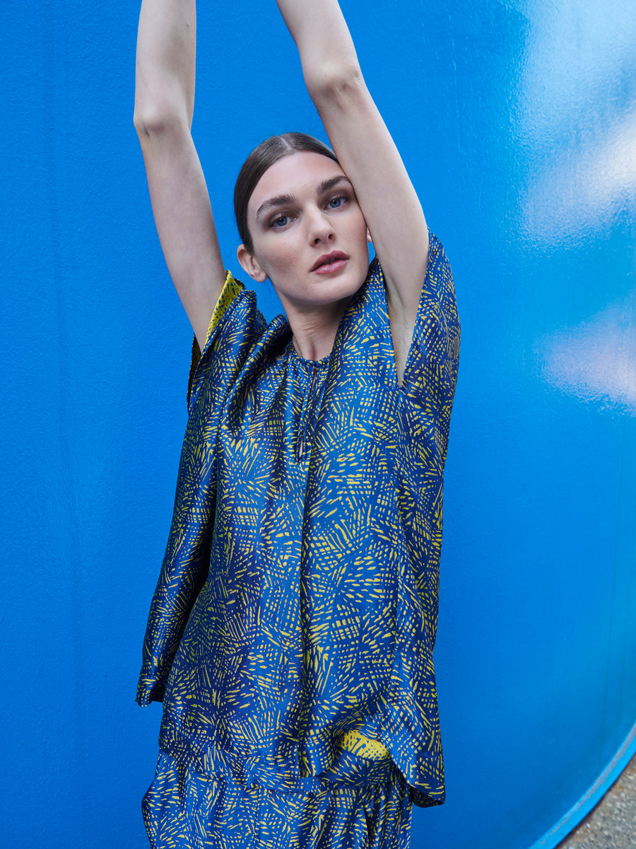 A long-haired person poses with arms raised against a bright blue background, wearing the sustainable Rae Poncho Top by Zero + Maria Cornejo. The outfit showcases vibrant blue and yellow jacquard patterns, beautifully crafted in New York.