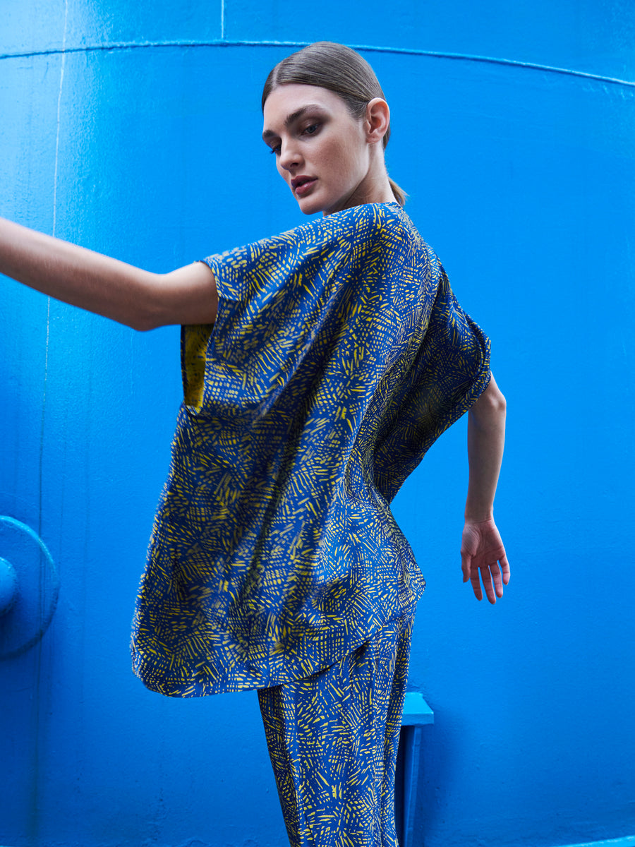 In a striking blue and yellow pattern, the Rae Poncho Top and matching pants by Zero + Maria Cornejo, both sustainably made in New York with a loose fit, are modeled dynamically against a vivid blue background. The person's hair is tied back as they glance to the side.