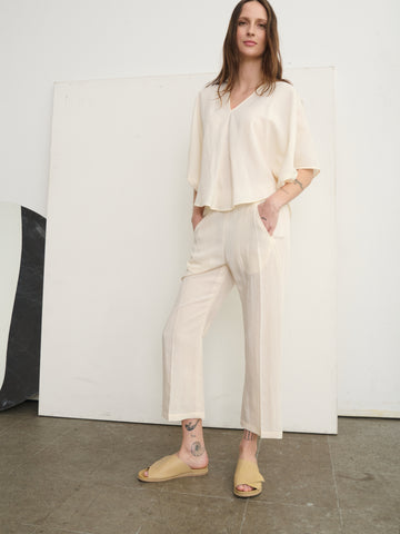 Against a plain backdrop, a person wears an airy top and Zero + Maria Cornejo's Eko Pant, featuring technical woven fabric and an elastic waist. They have long hair, wear slide sandals, have tattoos on their right ankle, and place their hands in their pockets.