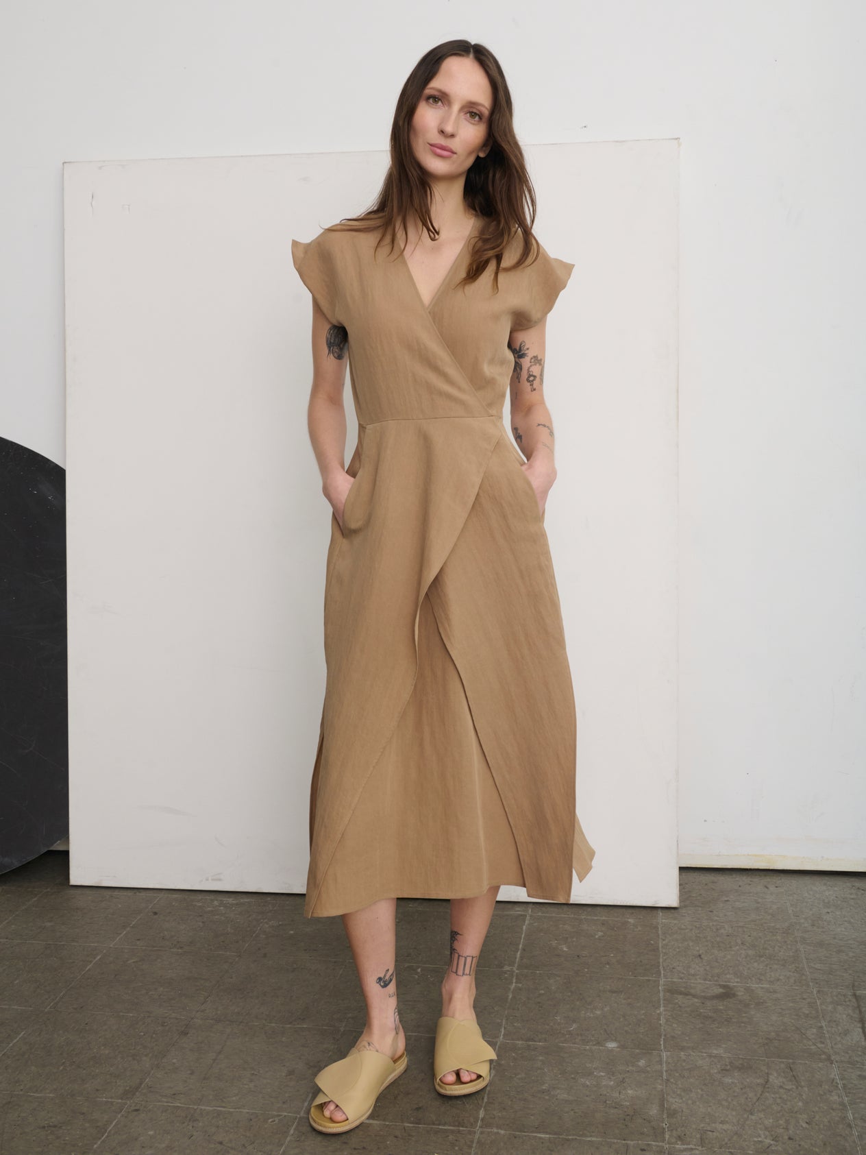 A person stands on stairs against an orange wall, wearing a vibrant orange Mido Dress by Zero + Maria Cornejo and white sandals. They gaze ahead confidently, partially leaning on the railing for support.