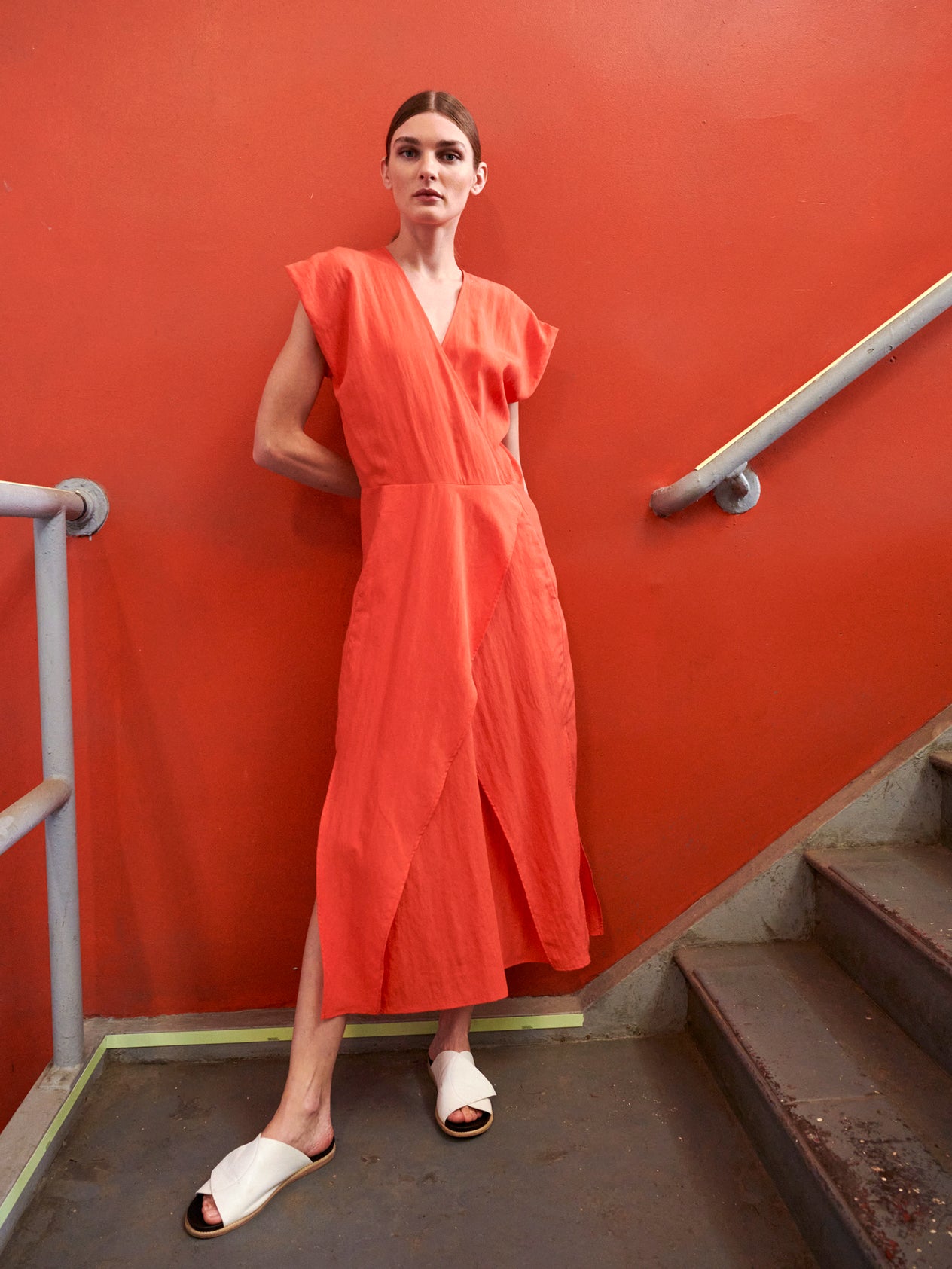 A person stands on stairs against an orange wall, wearing a vibrant orange Mido Dress by Zero + Maria Cornejo and white sandals. They gaze ahead confidently, partially leaning on the railing for support.