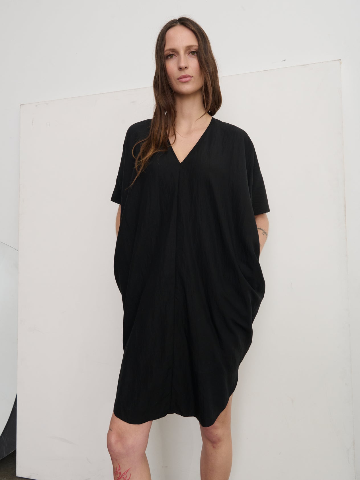 A person stands against a concrete wall wearing the oversized, lightweight Circe Dress by Zero + Maria Cornejo in orange, featuring a V-neck. Paired with black sandals, they strike a casual pose with one leg slightly bent and arms partially outstretched.