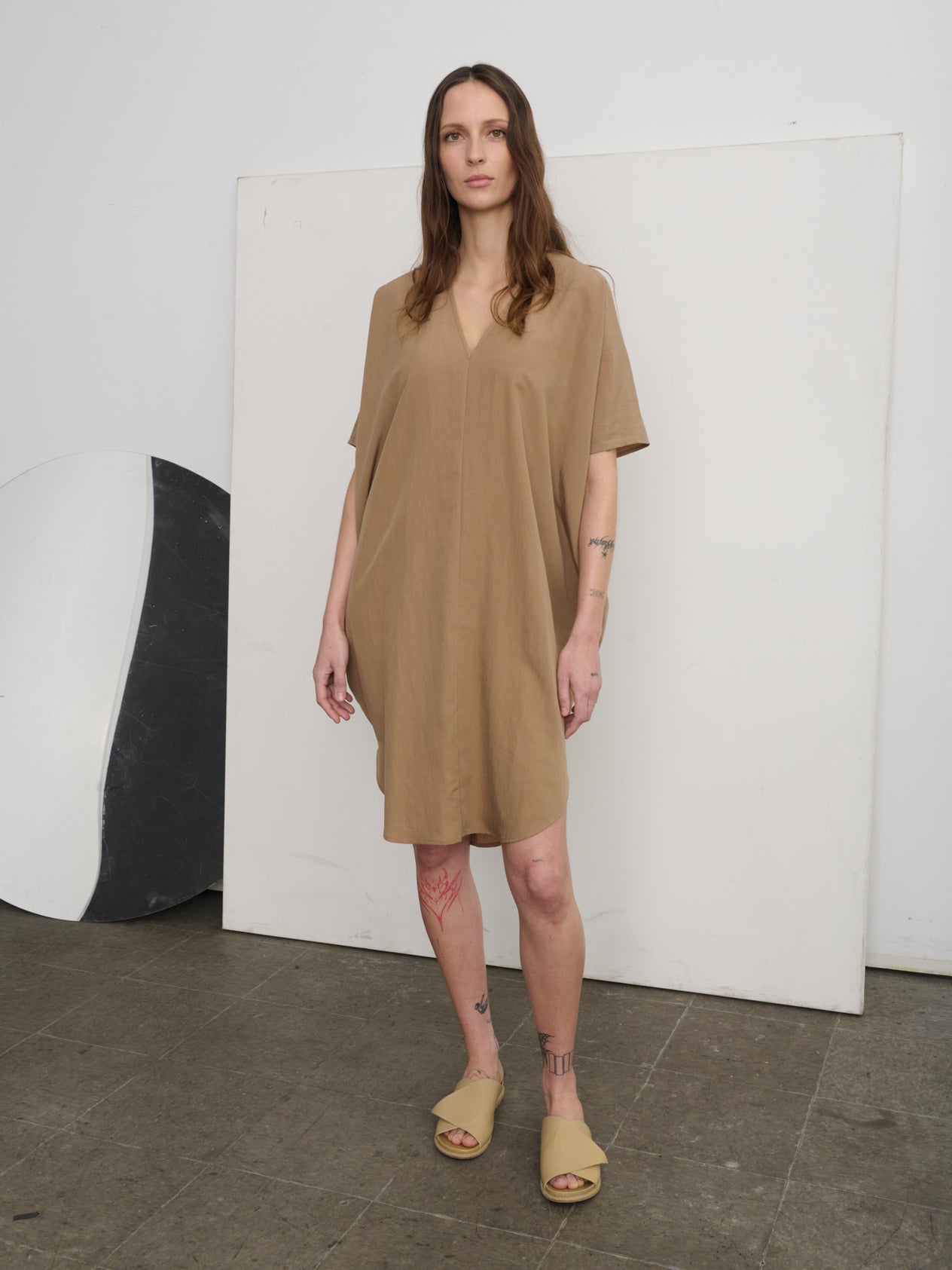 A person stands against a concrete wall wearing the oversized, lightweight Circe Dress by Zero + Maria Cornejo in orange, featuring a V-neck. Paired with black sandals, they strike a casual pose with one leg slightly bent and arms partially outstretched.