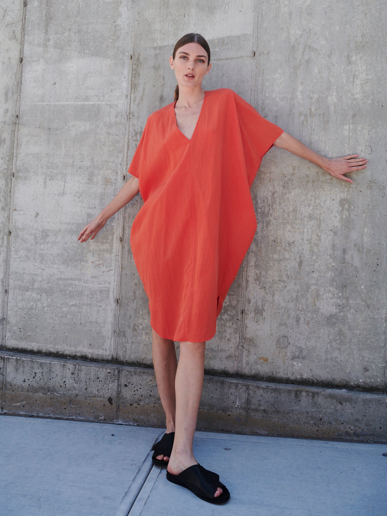 A person stands against a concrete wall wearing the oversized, lightweight Circe Dress by Zero + Maria Cornejo in orange, featuring a V-neck. Paired with black sandals, they strike a casual pose with one leg slightly bent and arms partially outstretched.