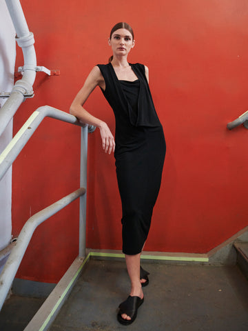 A relaxed individual in the Cowl Dress by Zero + Maria Cornejo, paired with sandals, leans against a staircase railing in front of an orange wall. One foot is on a lower step, and pipes can be seen in the top left corner.