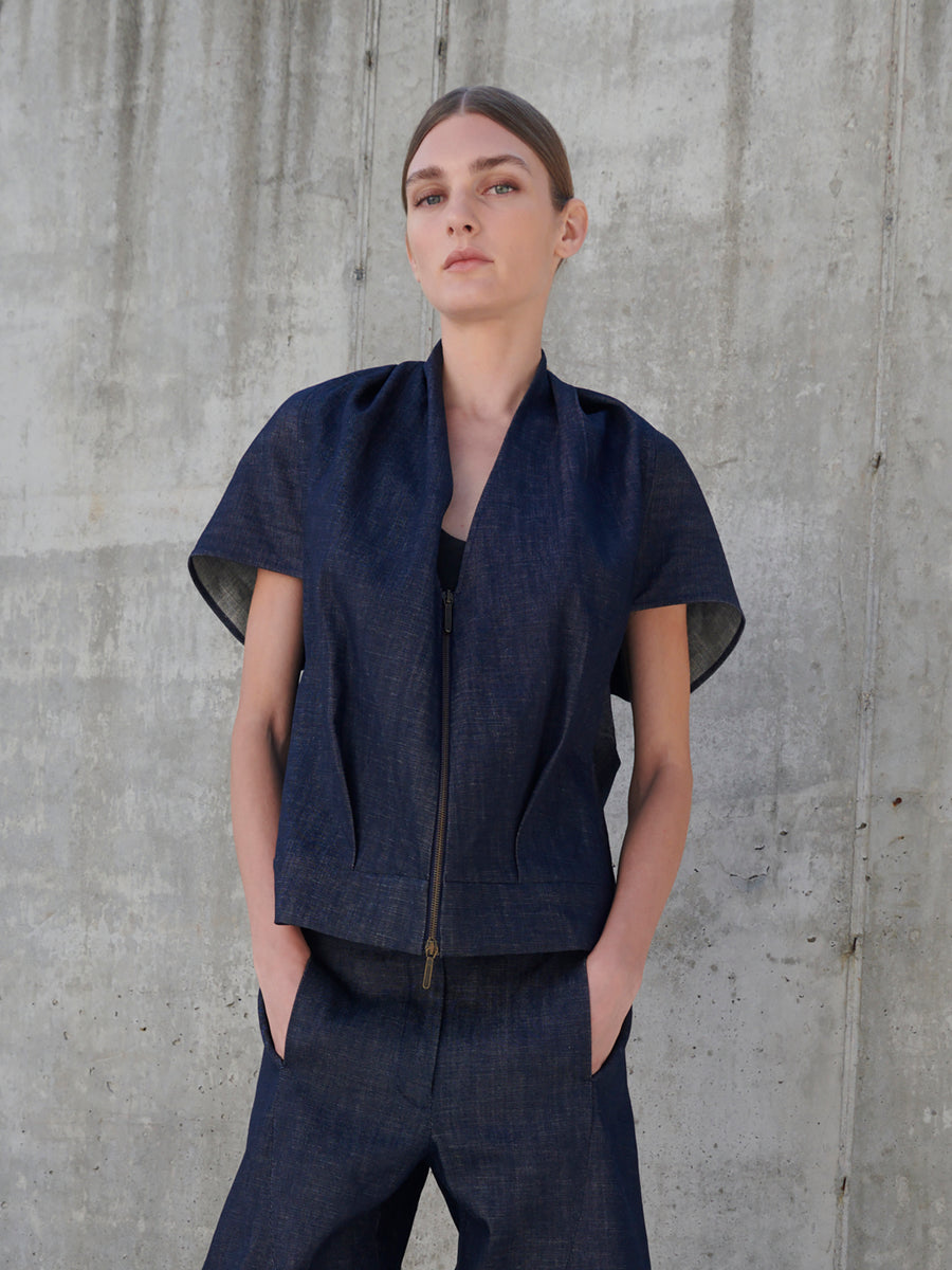 Against a concrete wall, the person wears Zero + Maria Cornejo's dark Maki Shrug and matching pants. Hands in pockets, they gaze upward with neatly tied-back hair, the outfit's pleated front adding elegance to their calm demeanor.