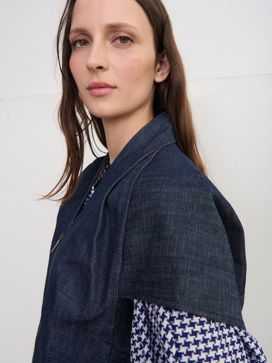 A person with long brown hair faces the camera, styled in a Maki Shrug by Zero + Maria Cornejo over a patterned shirt. They stand against a plain white wall, emphasizing their style.