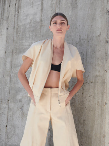 A person with straight hair stands against a concrete wall, wearing a Maki Shrug by Zero + Maria Cornejo. The beige cocoon silhouette ensemble includes pleated front details with a loose, short-sleeve cardigan and high-waisted pants over a black top, hands casually in pockets.