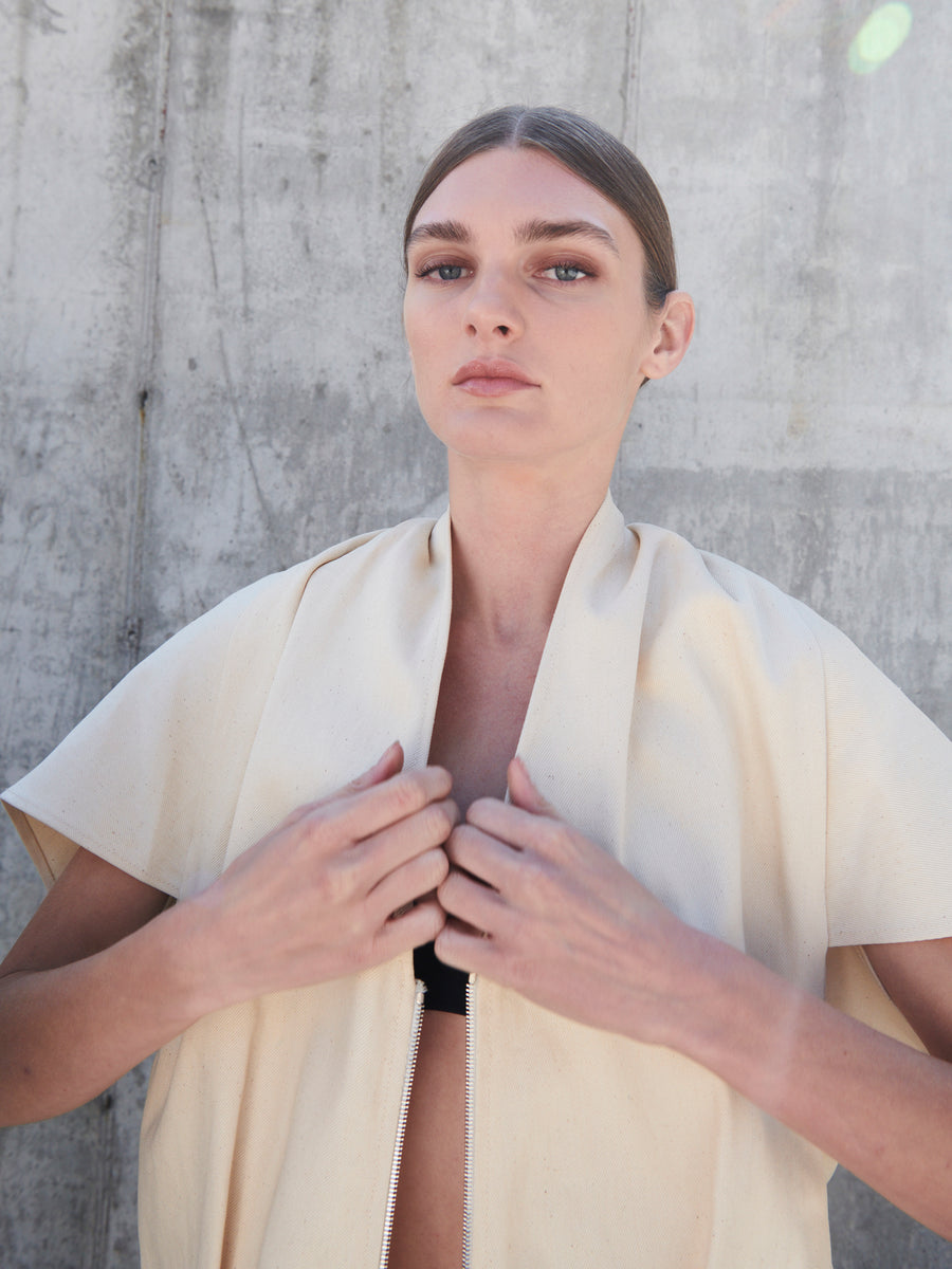 A person with long hair pulled back gazes confidently at the camera, wearing the Maki Shrug by Zero + Maria Cornejo. Standing against a concrete wall, the image conveys a minimalist and serene mood.