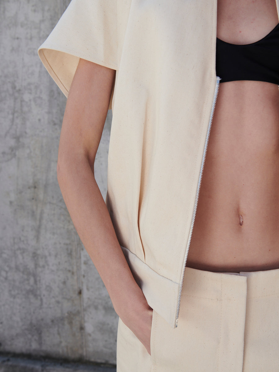 A person showcases the Maki Shrug by Zero + Maria Cornejo—a cream-colored, cocoon silhouette, short-sleeve jacket with pleated front detail—partially zipped over a black top. They stand with one hand in their pocket against a concrete wall, highlighting the clothing and midsection.