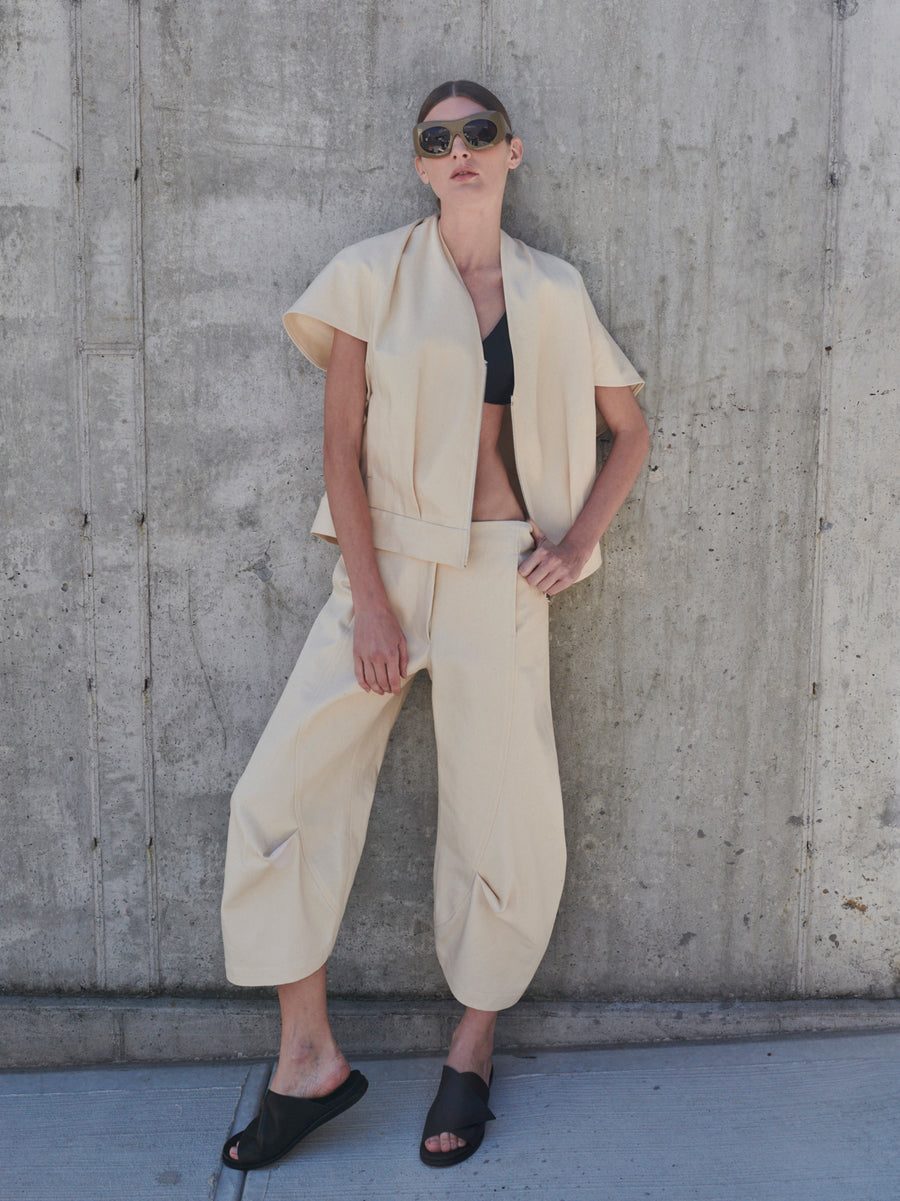 Dressed in zero + Maria Cornejo's Maki Shrug, along with beige loose pants and black sandals, a person confidently poses against a concrete wall, sunglasses on, highlighting a chic contemporary cocoon silhouette.