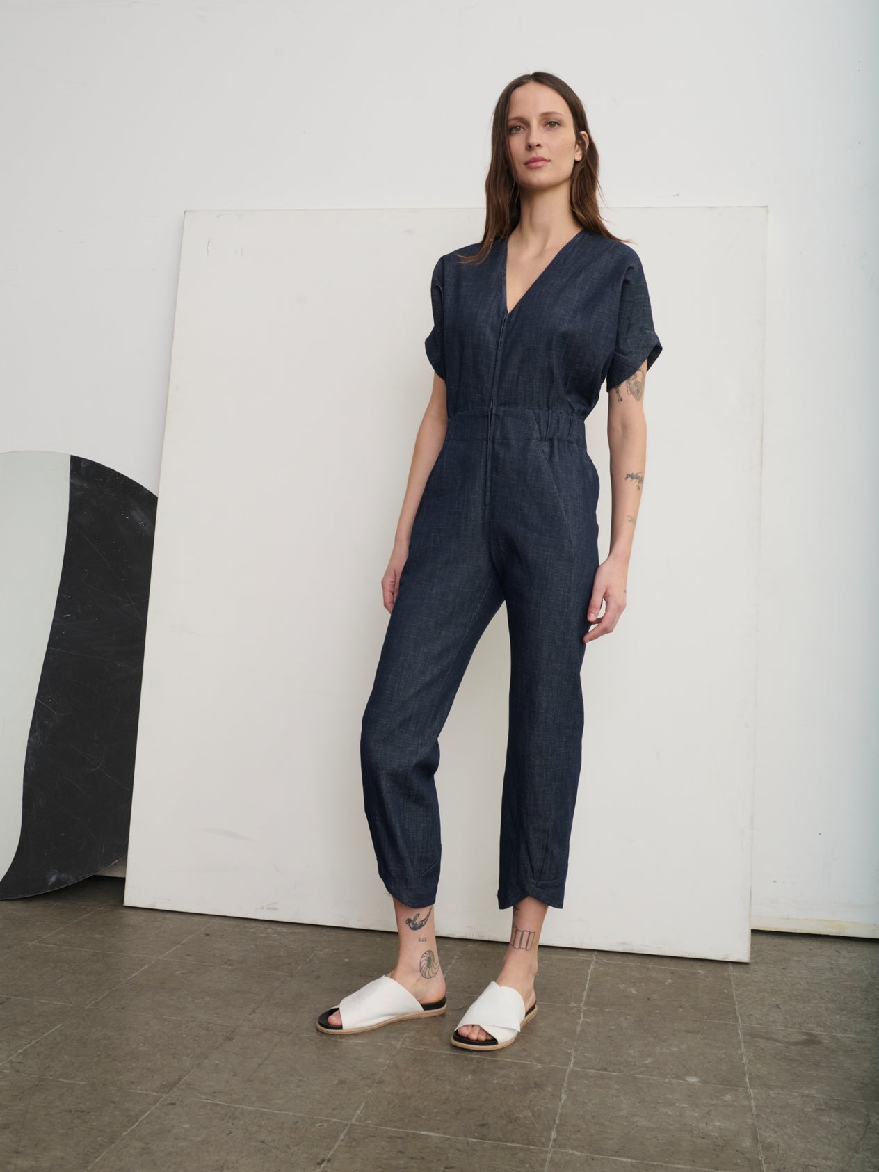 A person in a cream Aissa V-Neck Jumpsuit by Zero + Maria Cornejo and white sandals stands on a staircase with a bright orange wall, one hand in pocket, looking forward.