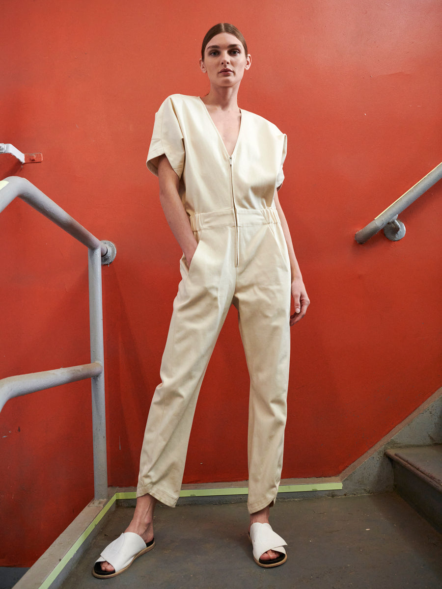 A person in a cream Aissa V-Neck Jumpsuit by Zero + Maria Cornejo and white sandals stands on a staircase with a bright orange wall, one hand in pocket, looking forward.