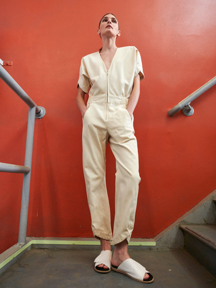 On a staircase, a person in the Aissa V-Neck Jumpsuit by Zero + Maria Cornejo and white slippers is framed by an orange wall and gray railings, gazing upward with a contemplative expression.