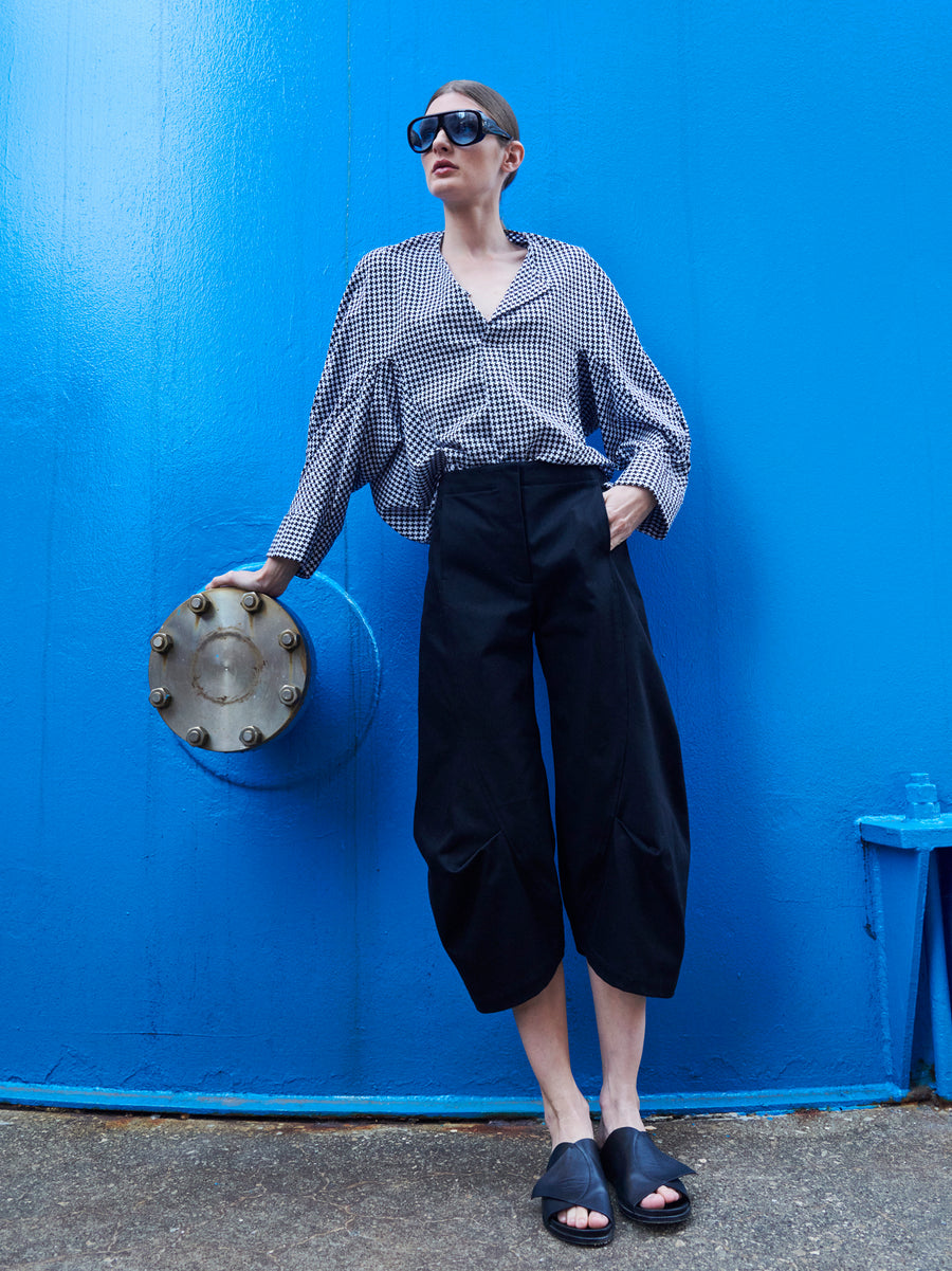 A person wearing a black-and-white checkered shirt, Beetle Culotte by Zero + Maria Cornejo, sunglasses, and sandals stands beside a vibrant blue industrial structure with a round metal bolt panel. Their hand rests on the metal for a stylish urban scene.