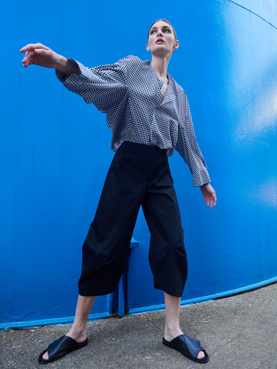 A person in a checkered blouse and Beetle Culotte by Zero + Maria Cornejo poses dramatically against a bright blue wall. They wear black slide sandals, captured from a low angle with one arm slightly extended.