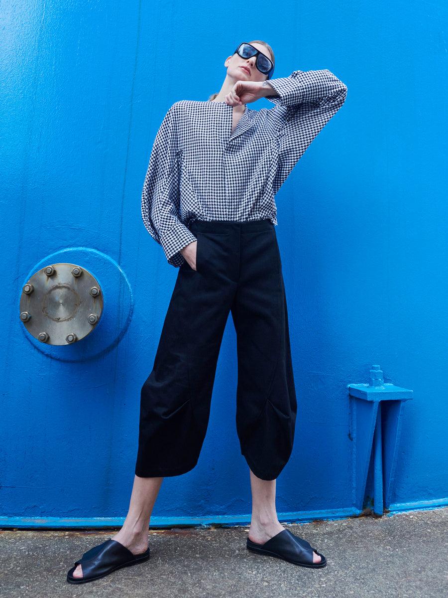 A confident person in sunglasses and a checkered shirt leans against a vibrant blue wall, wearing Zero + Maria Cornejo's Beetle Culotte black cropped pants and black slide sandals, with one hand in their pocket.