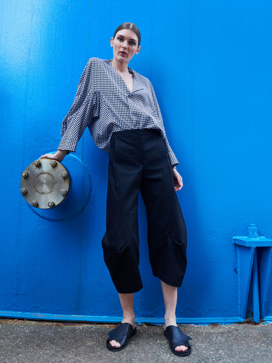 A person poses confidently against a vibrant blue wall, wearing a black and white checkered shirt made from GOTS-certified cotton, Beetle Culotte pants by Zero + Maria Cornejo, and black slide sandals. They are leaning on a circular metal fixture.