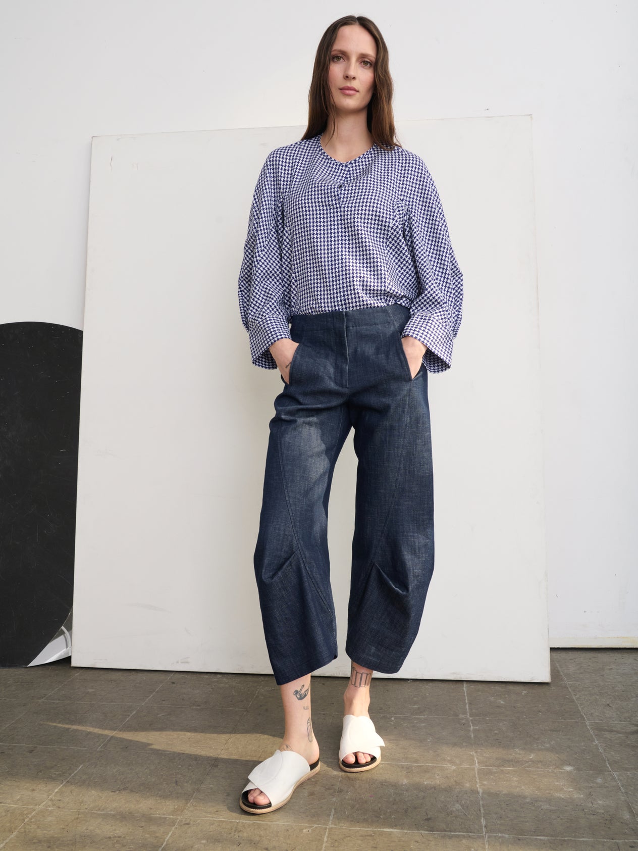 Against a concrete wall, a person stands confidently wearing the Beetle Culotte set by Zero + Maria Cornejo, made from GOTS-certified cotton. The outfit features a beige high-waisted pant and jacket over a black crop top, completed with black slide sandals. Hands are in pockets with feet apart.