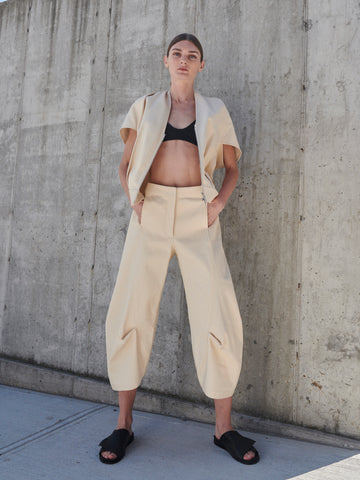 Against a concrete wall, a person stands confidently wearing the Beetle Culotte set by Zero + Maria Cornejo, made from GOTS-certified cotton. The outfit features a beige high-waisted pant and jacket over a black crop top, completed with black slide sandals. Hands are in pockets with feet apart.