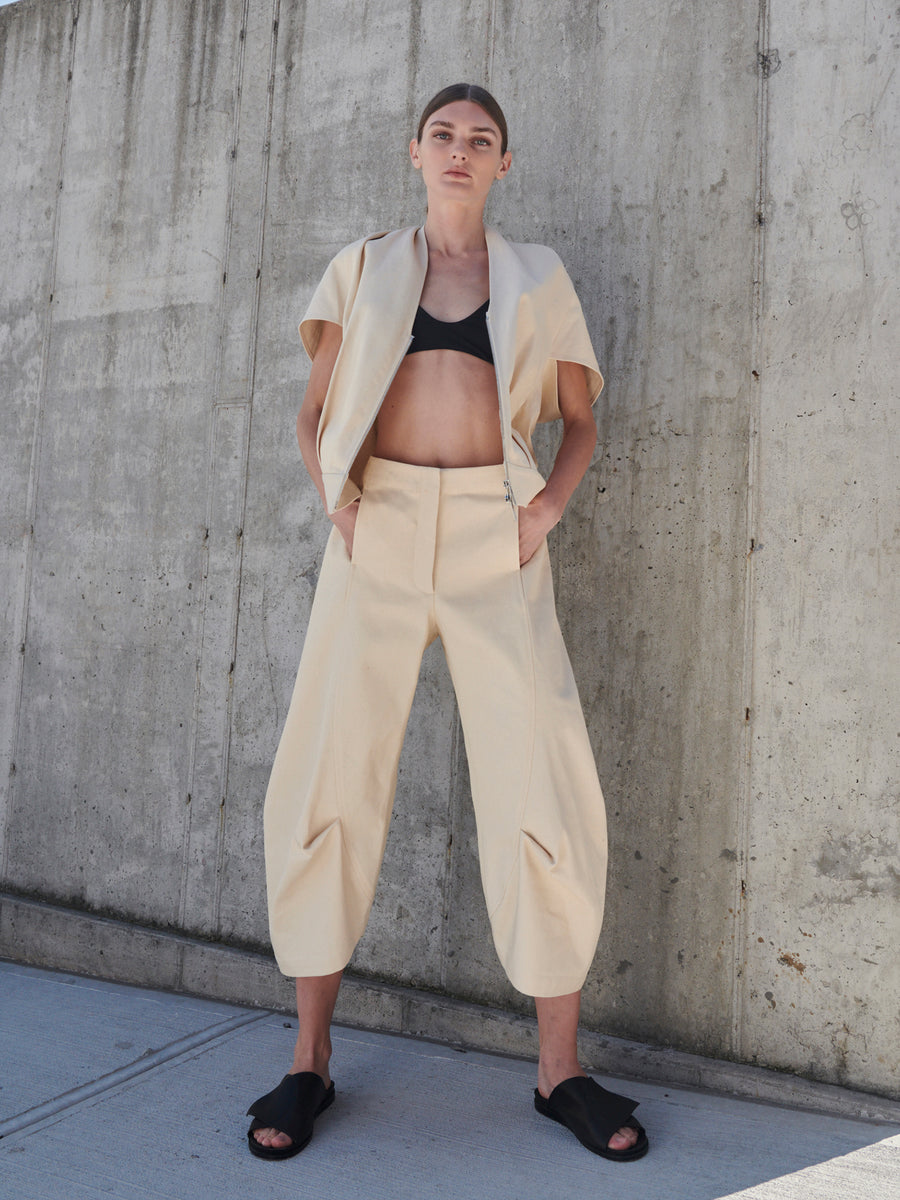 Against a concrete wall, a person stands confidently wearing the Beetle Culotte set by Zero + Maria Cornejo, made from GOTS-certified cotton. The outfit features a beige high-waisted pant and jacket over a black crop top, completed with black slide sandals. Hands are in pockets with feet apart.