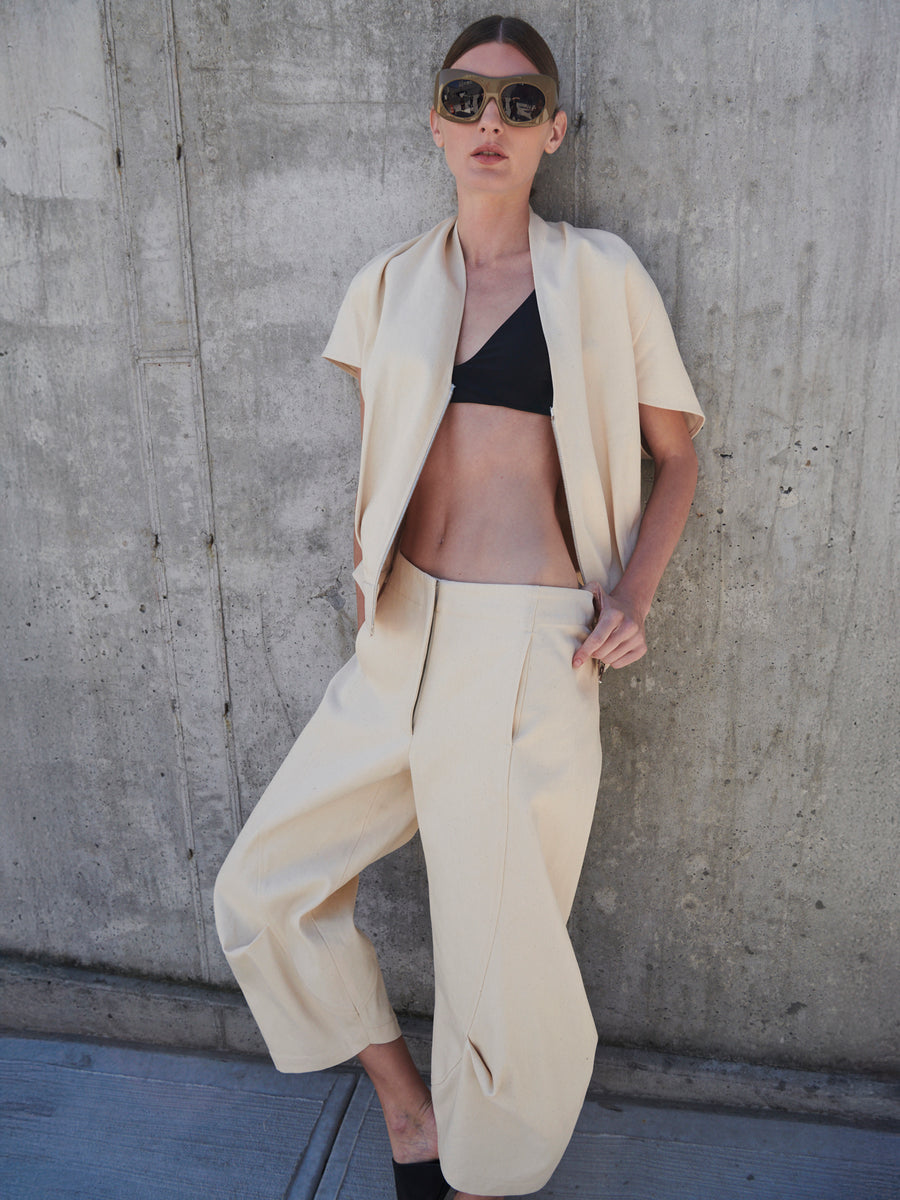 A woman wearing a black crop top and the Beetle Culotte by Zero + Maria Cornejo, made from GOTS-certified cotton, a cream-colored jacket, and sunglasses leans against a concrete wall with her hands in her pockets.