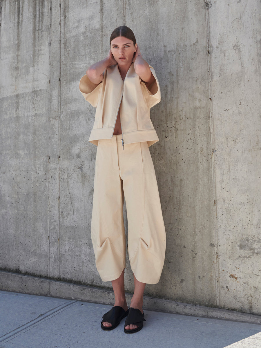 A person stands against a concrete wall, wearing Zero + Maria Cornejo's Beetle Culotte in cream and an open GOTS-certified cotton jacket, paired with black sandals. Their long hair is tied back as they raise their hands near their neck.