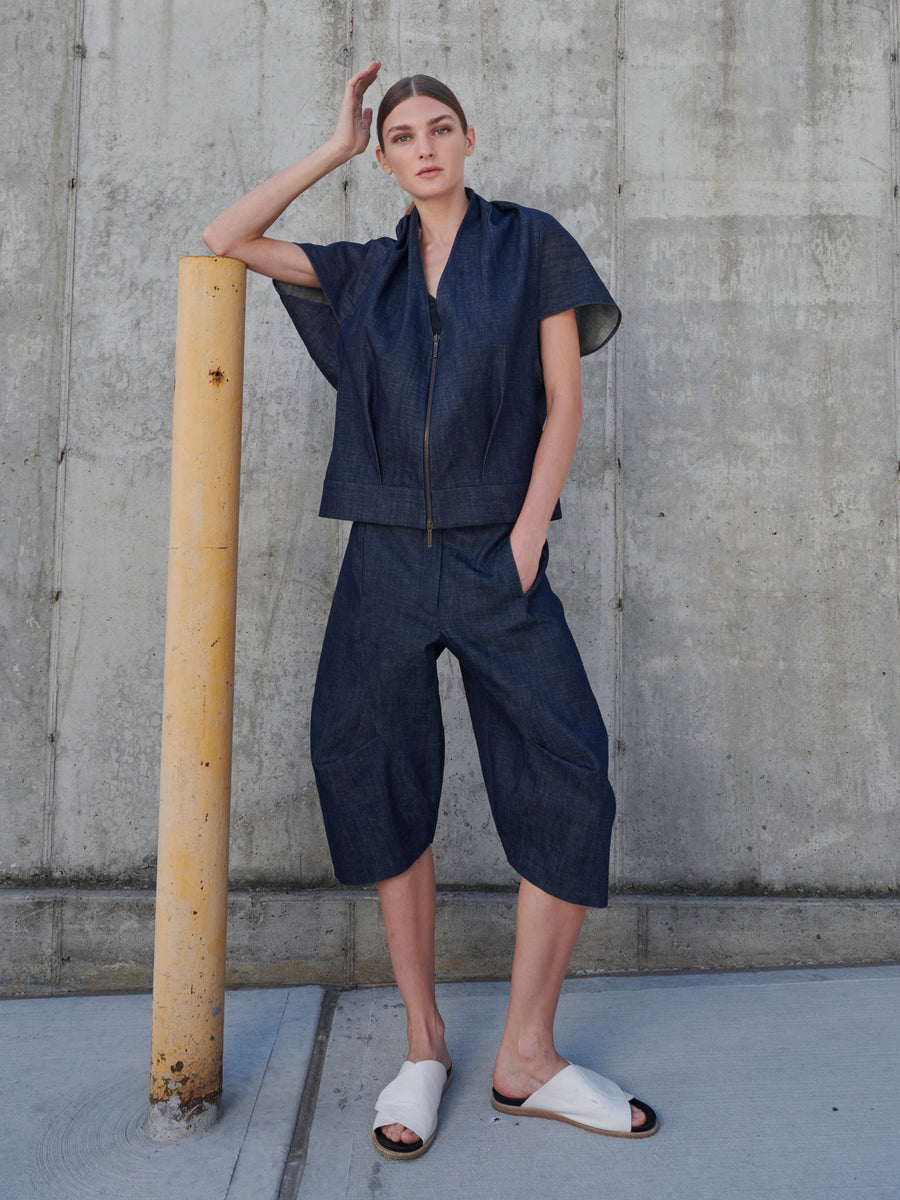 A person leans against a concrete wall, resting on a yellow pole. They're dressed in the Beetle Short and a denim zip-up jacket, with their left hand tucked into a pocket. They complete the look with white slide sandals. Brand: Zero + Maria Cornejo.